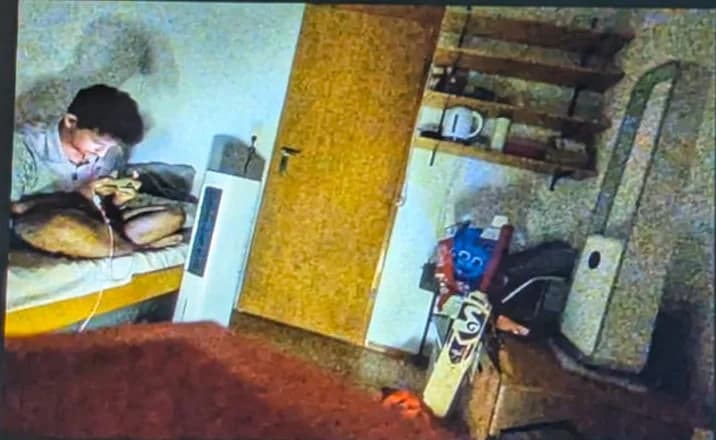 Orio's camera view of a bedroom: a person sitting on a bed, a door, shelves and a cricket bat leaning against a box